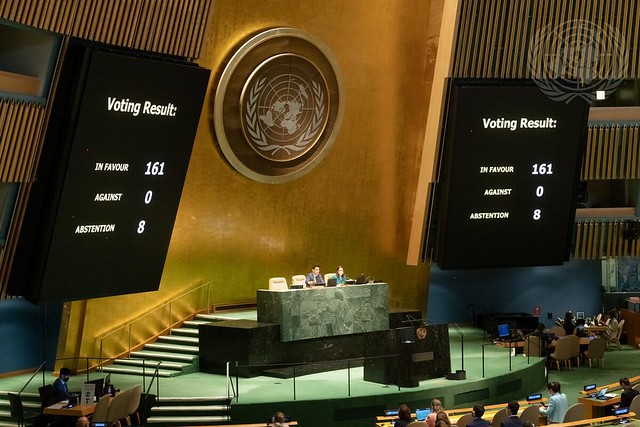 A photo of the UN amphitheater stage with two large black screens on either side displaying voting results.  