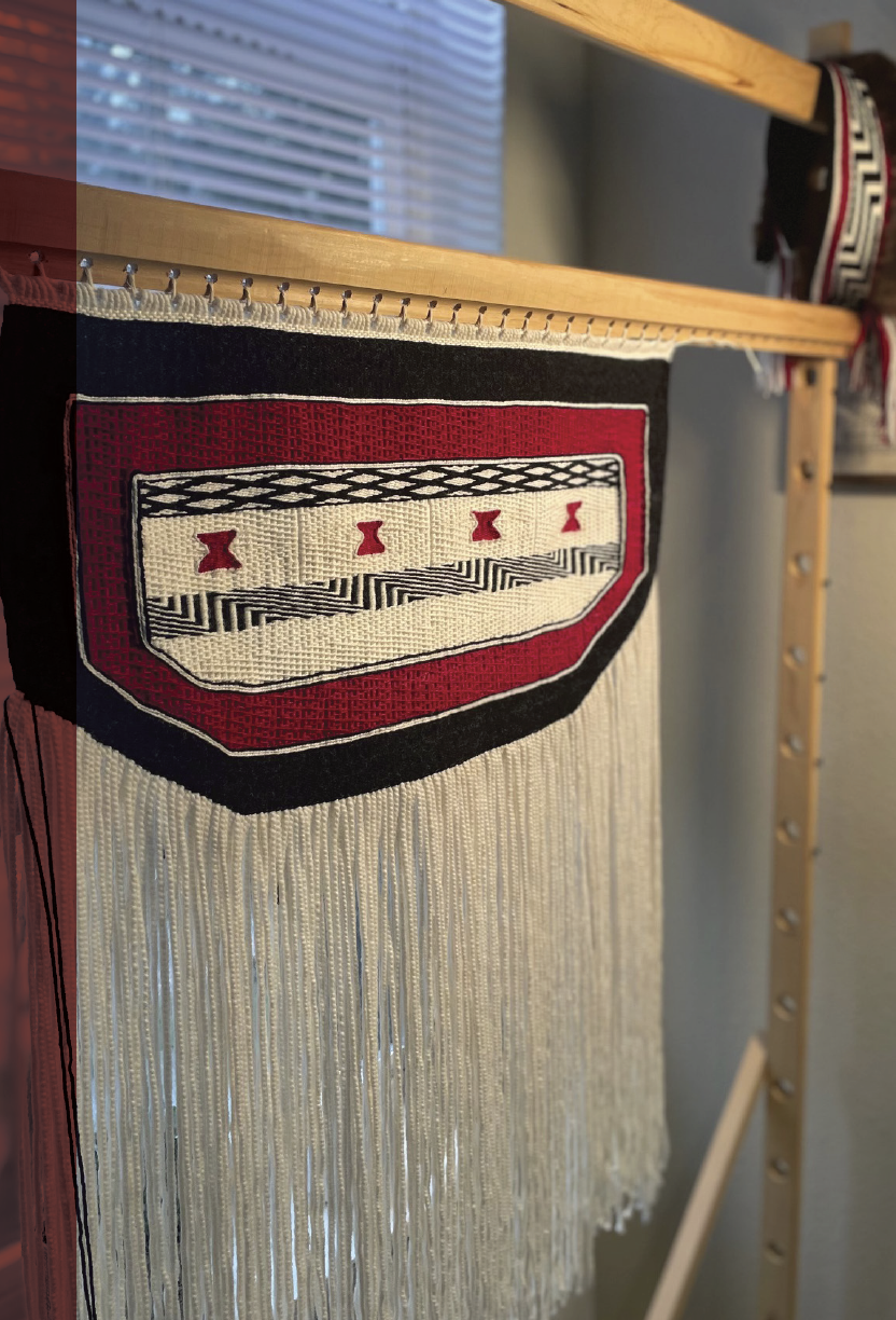"Red, black, and white Ravenstail woven apron hanging on a wooden loom"