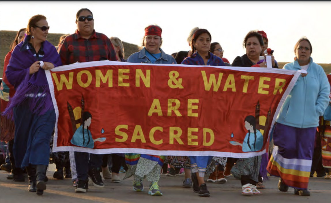 "A line of native women walking towards the camera, holding a red banner that says 'Women and Water are Sacred'""