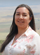 Keely Linton standing outside, she has long brown hair and is smiling with her teeth, wearing a white and pink floral shirt. 