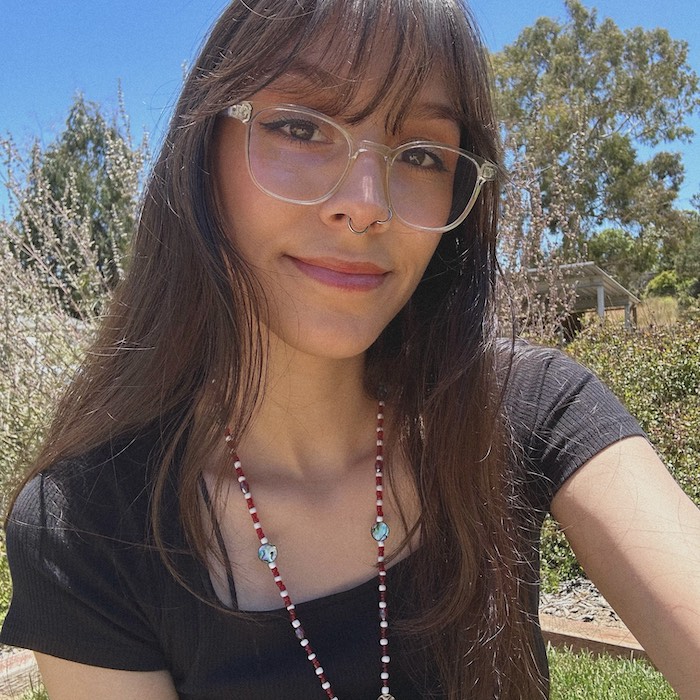 Brianna sitting outside in the desert, wearing clear glasses, a long delicate necklace and she has long brown hair with bangs. 